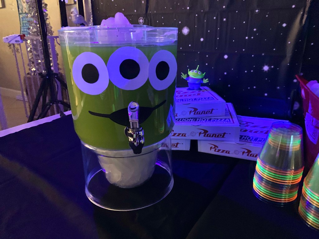 Green alien-themed drink dispenser with glowing green punch beside Pizza Planet boxes.