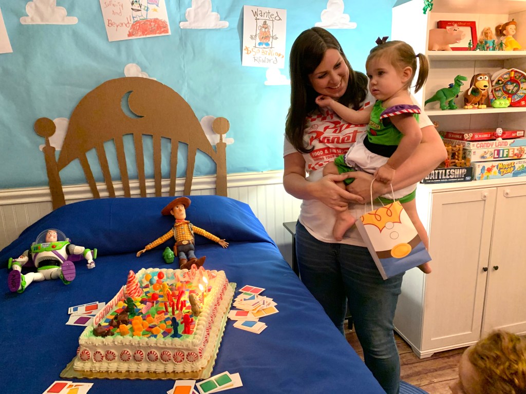 Mother holds toddler in Buzz Lightyear dress beside a Candy Land birthday cake on a bed with Toy Story dolls.