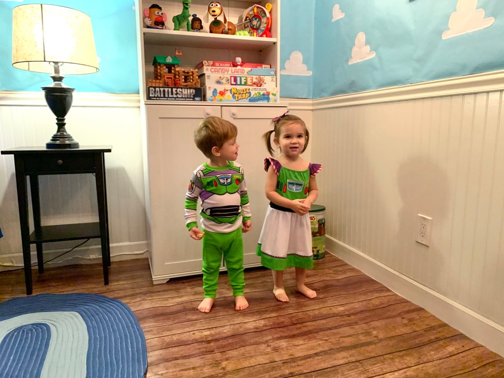 Two toddlers wearing Buzz Lightyear costumes stand side by side in a Toy Story-decorated room.