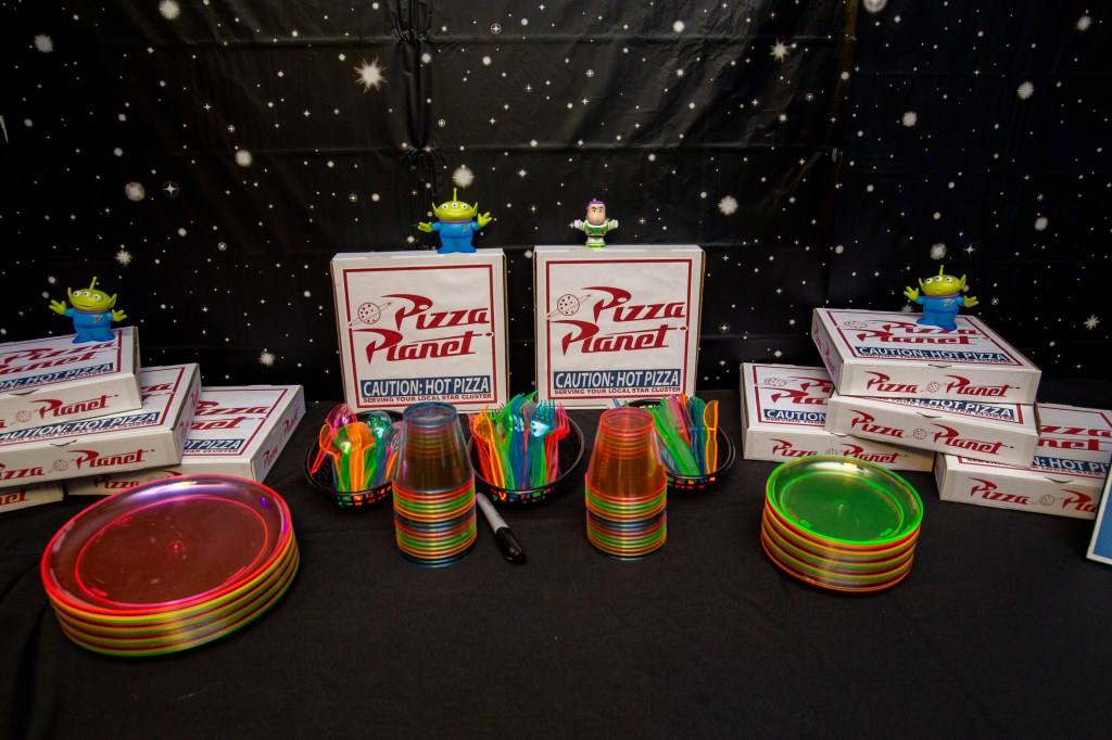 Pizza Planet-themed table setup with stacked pizza boxes, glow utensils, and alien figurines, before food service begins.