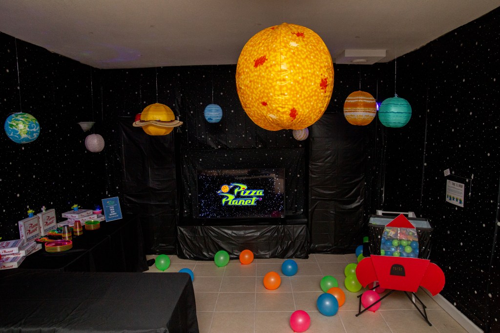 A Toy Story-themed party room decorated like Pizza Planet with hanging planets, a Pizza Planet logo on the TV, black starry walls, balloons, and a red rocket-shaped prize machine.