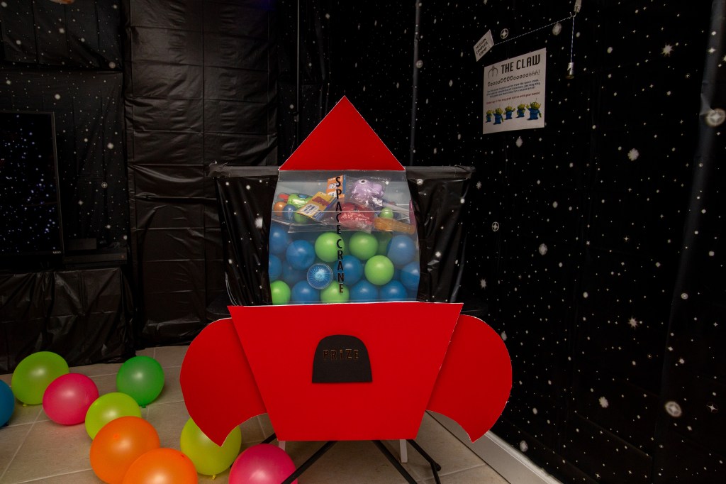 A red cardboard rocket ship labeled “Prize” holds a bin of colorful balls and small toys, recreating the iconic Pizza Planet claw machine from Toy Story.