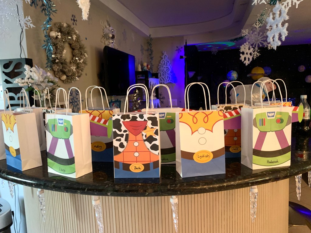 A row of white favor bags decorated with Toy Story character outfits such as Buzz Lightyear, Woody, and Bo Peep, each labeled with a guest’s name.