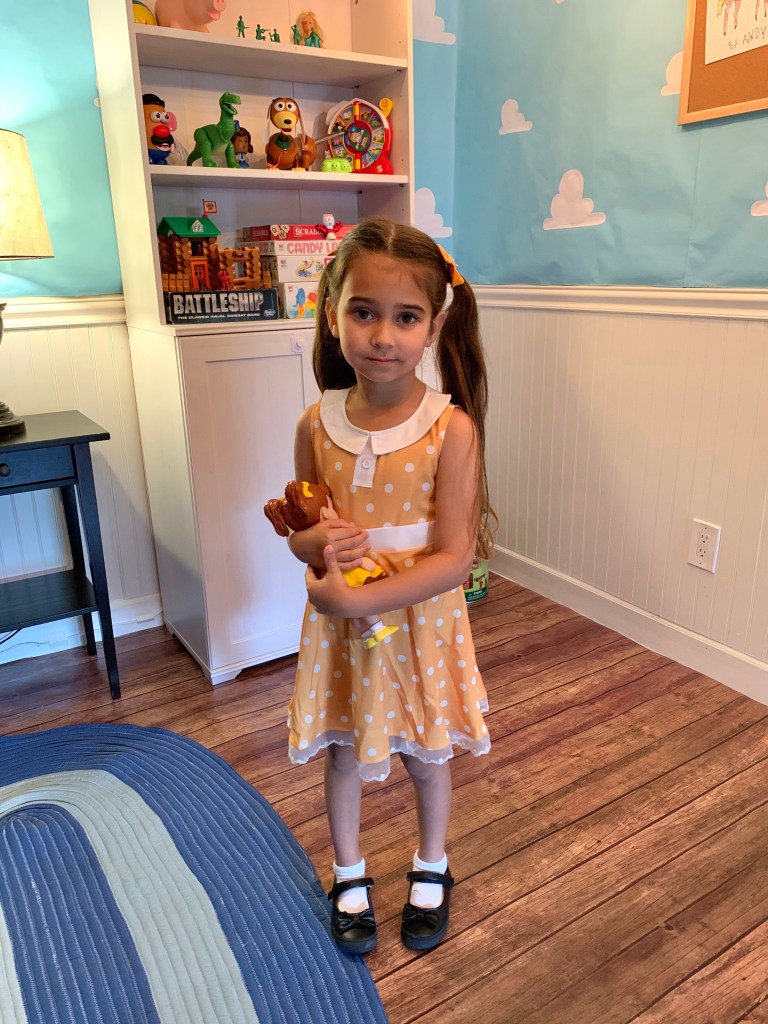Young girl wearing a yellow Gabby Gabby dress stands in a Toy Story-themed bedroom holding a Gabby Gabby doll.