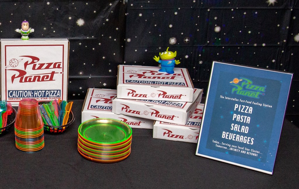 Toy Story-themed Pizza Planet table display with custom pizza boxes, blacklight-reactive plates and utensils, and a printed Pizza Planet menu sign.