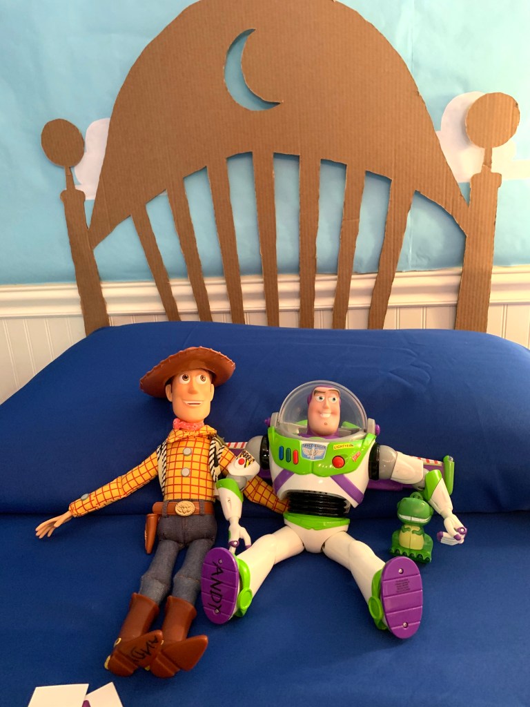 Woody and Buzz Lightyear dolls on blue bedding in front of handmade cardboard headboard
