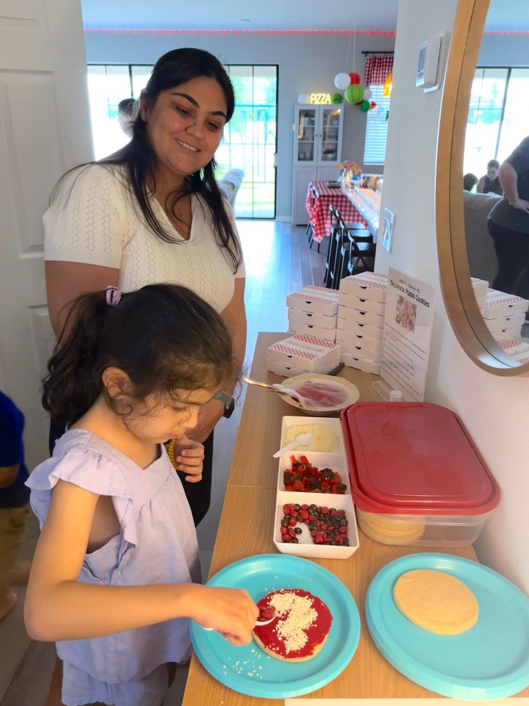 A young girl decorates a sugar cookie with red frosting and white sprinkles to look like a pizza, while a woman in a white top smiles beside her. Bowls of toppings and mini pizza boxes are arranged on the table.