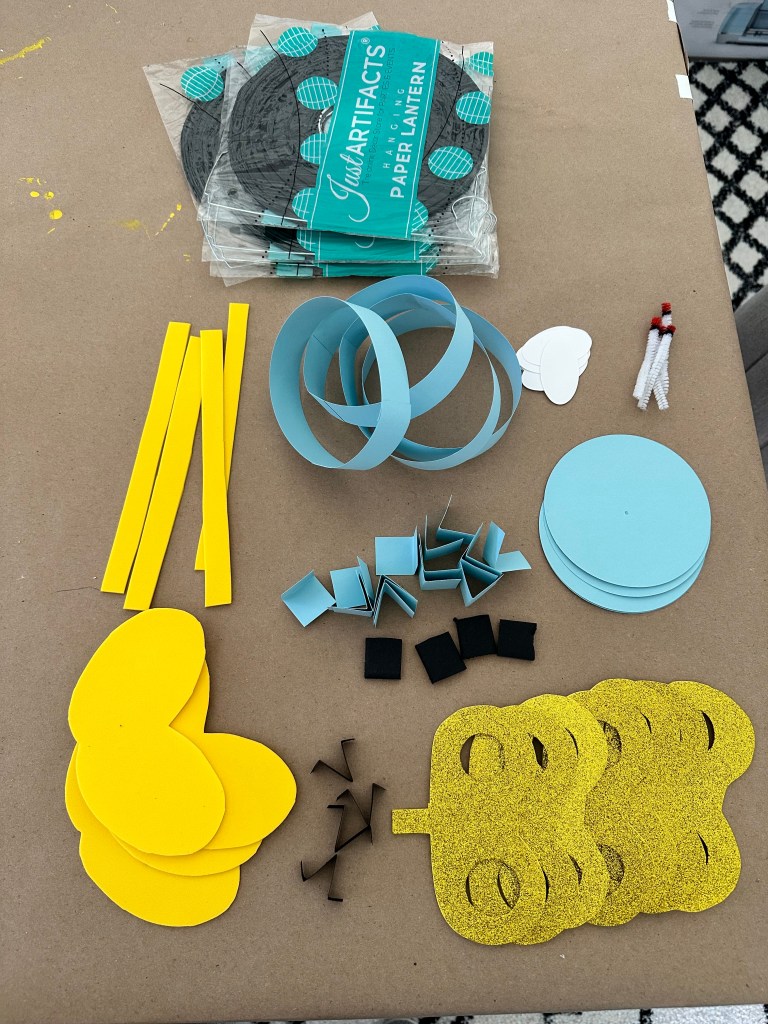 DIY Bob-omb tutorial materials laid out, including black paper lanterns, yellow foam, blue cardstock, and glittery gold shapes.
