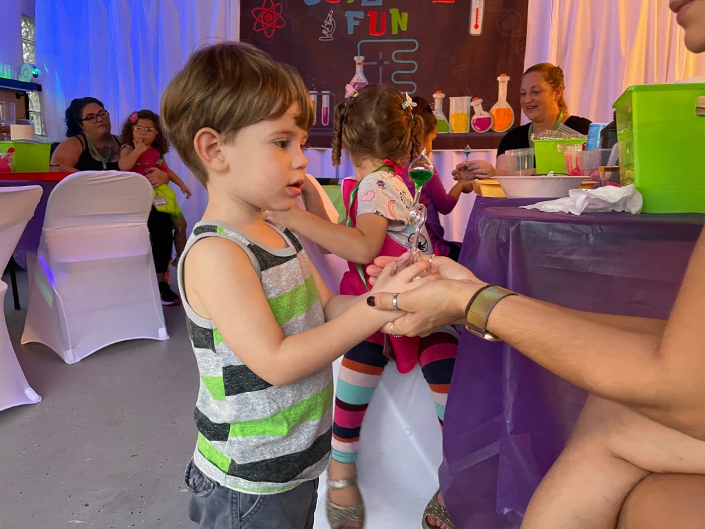 Young boy in a striped tank top looks at a clear hand boiler being handed to him in a science activity setting.