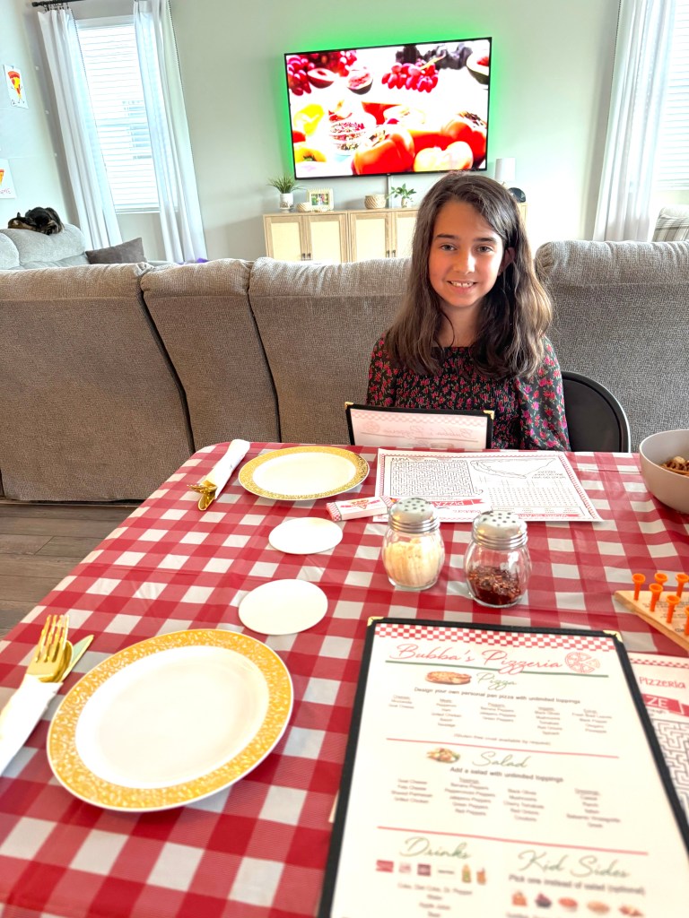 Elsa sitting at the freshly set table at Bubba’s Pizzeria, holding her menu and smiling, with a red-and-white checkered tablecloth and restaurant props.
