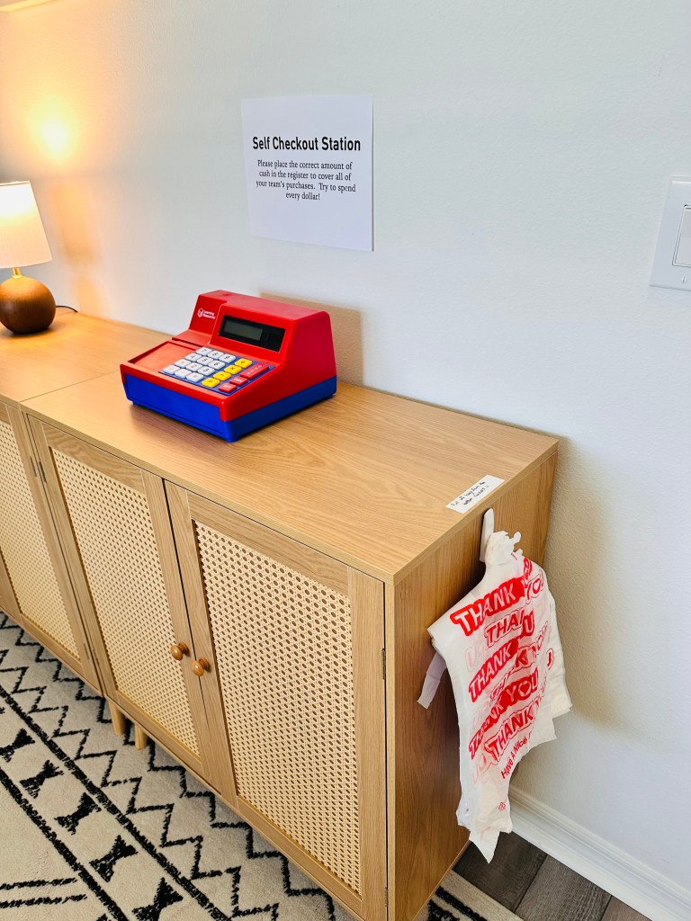A simple self checkout station with a toy cash register, “Self Checkout” sign, and plastic thank-you bags hanging on the side.