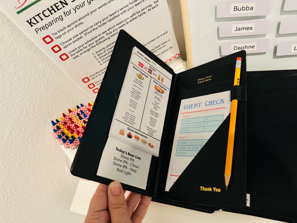 Close-up of a black restaurant-style guest check folder holding a printed Bubba’s Pizzeria menu, a guest order slip, and a yellow pencil, with crayon packs and a kitchen station sign visible in the background.