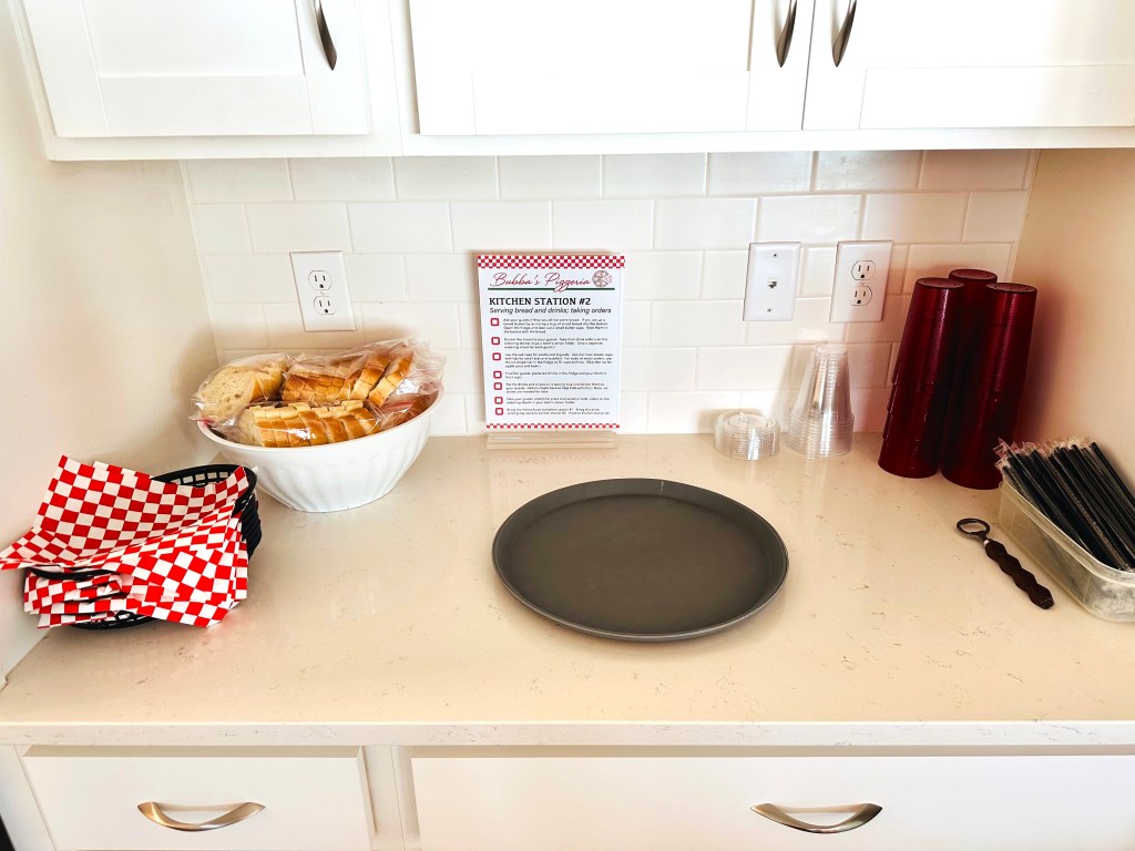 Kitchen Station 2 setup at Bubba’s Pizzeria party, showing bread, drink cups, tumblers, serving tray, straws, and instructional sign.