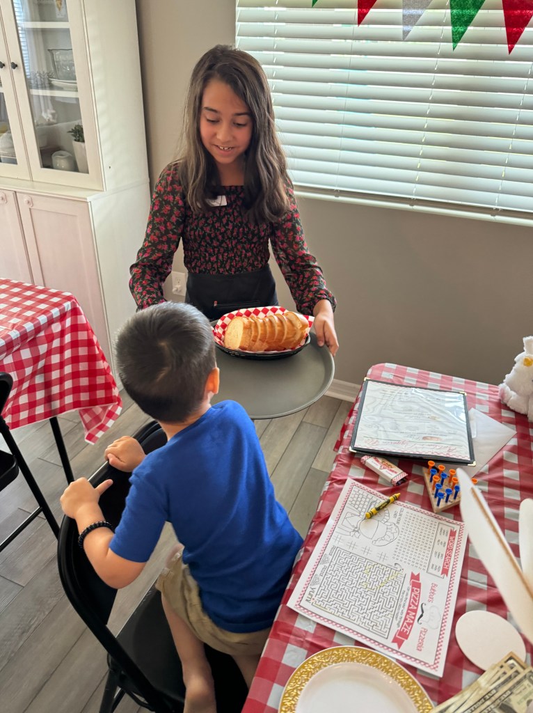 Child server delivering bread basket to seated guests at Bubba’s Pizzeria party, with activity placemats and crayons on the table.