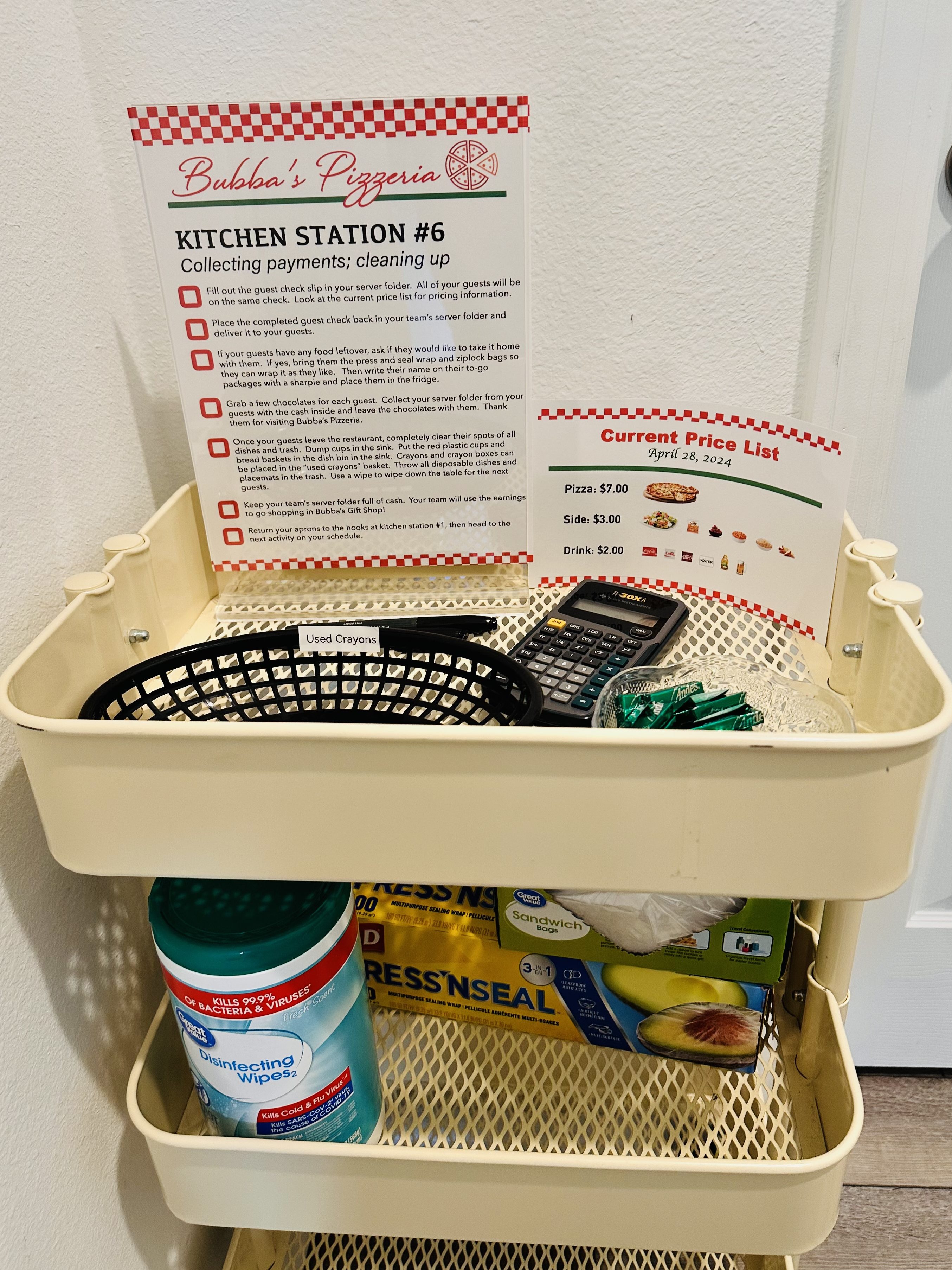 A beige rolling cart with instructions for Kitchen Station #6 at Bubba’s Pizzeria. The top shelf contains a sign with step-by-step instructions, a price list, calculator, black basket labeled “Used Crayons,” a bowl of Andes mints, and Sharpie markers. Lower shelves hold disinfecting wipes, press and seal wrap, and sandwich bags.