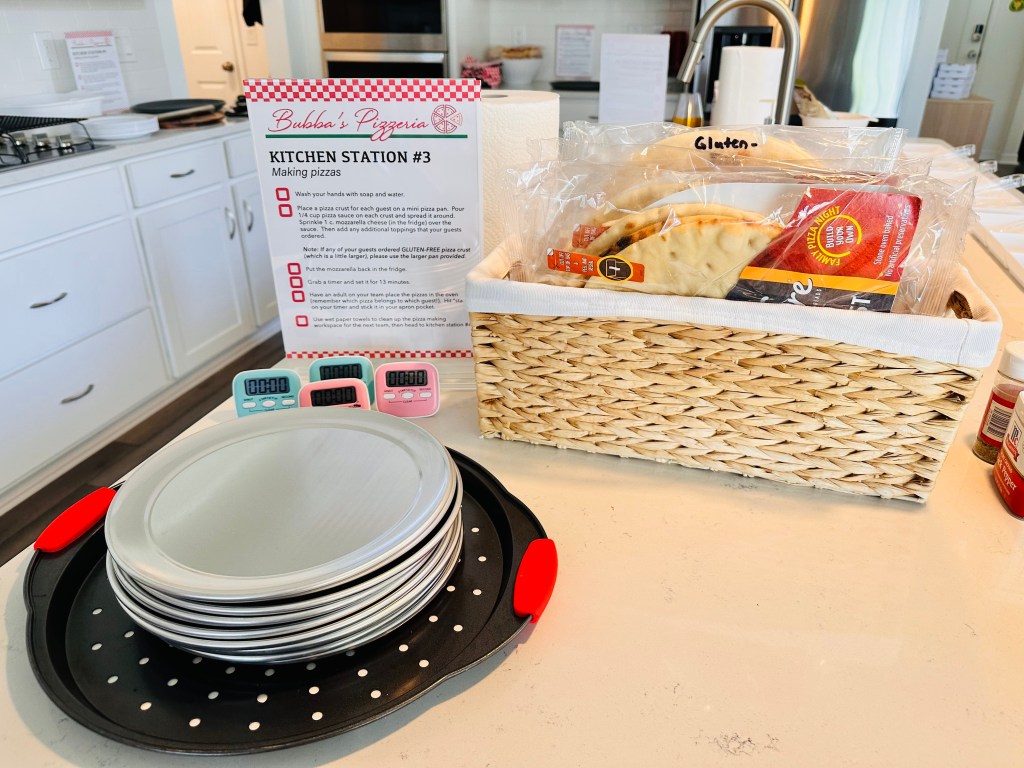 Countertop setup with plates, timers, and packaged pizza crusts next to a printed instruction sign labeled “Bubba’s Pizzeria Kitchen Station #3.”