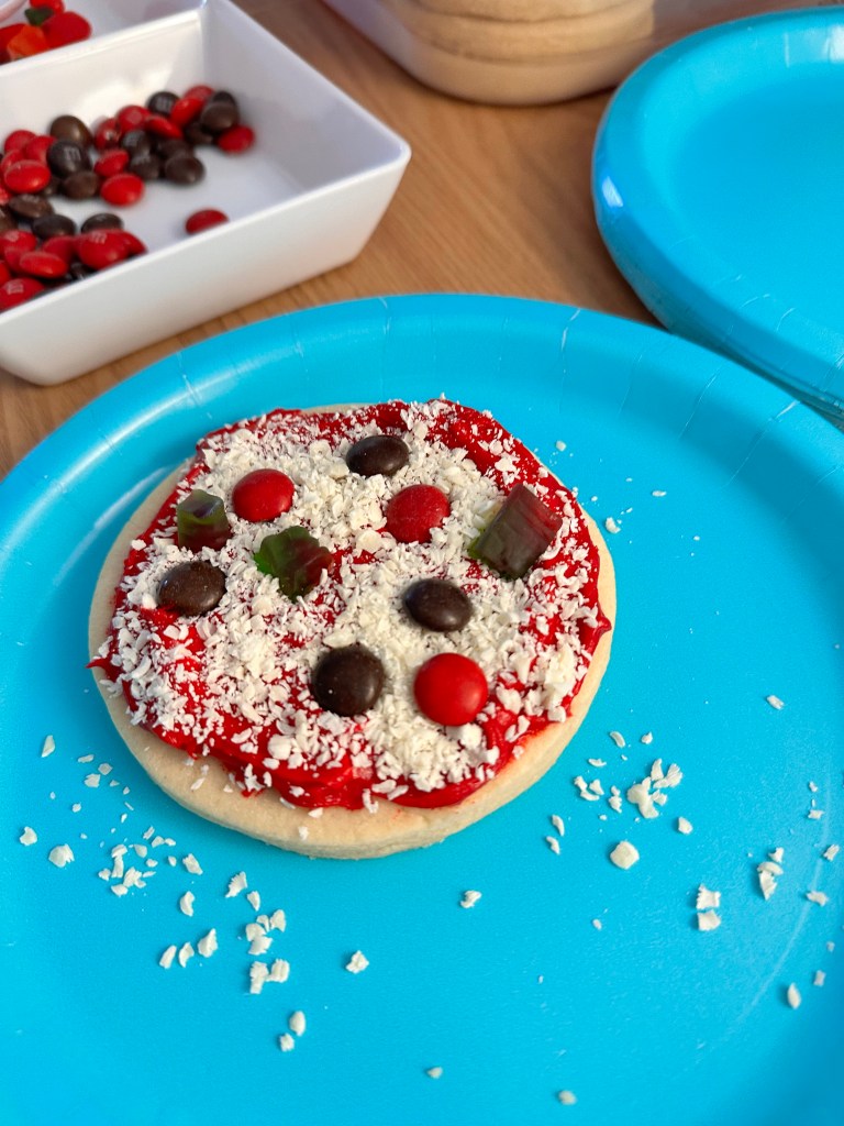 A sugar cookie decorated to look like a mini pizza, topped with red frosting “sauce,” shredded white chocolate “cheese,” and candy toppings like M&Ms and gummy pieces, sitting on a bright blue plate.