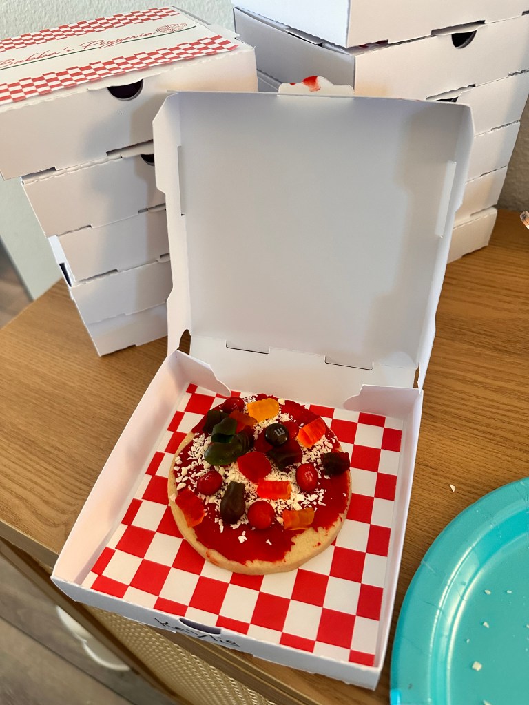 A decorated sugar cookie styled as a pizza, placed inside a mini pizza box with a red-and-white checkered liner. The cookie is topped with red frosting, shredded white chocolate, and colorful candy “toppings.”
