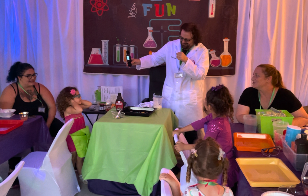 Man in a lab coat holds a decibel meter toward children in a decorated science lab party room as they prepare to scream.