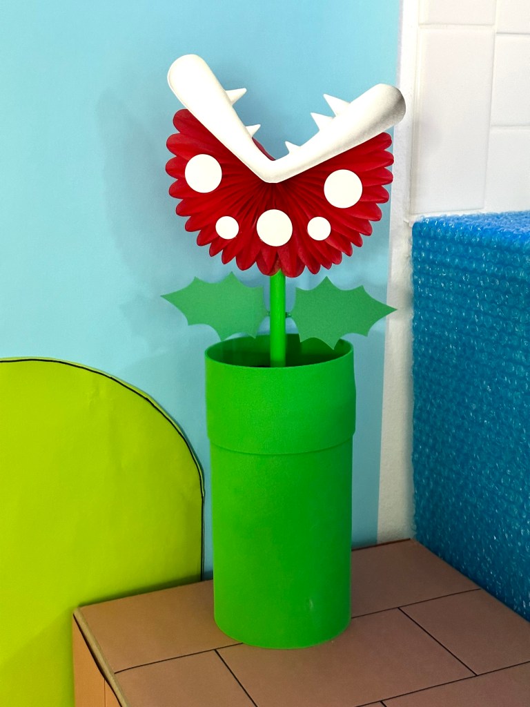 Close-up of a handmade Piranha Plant made from red paper fan, white teeth, and green stem inside a green pipe for Super Mario party decor.