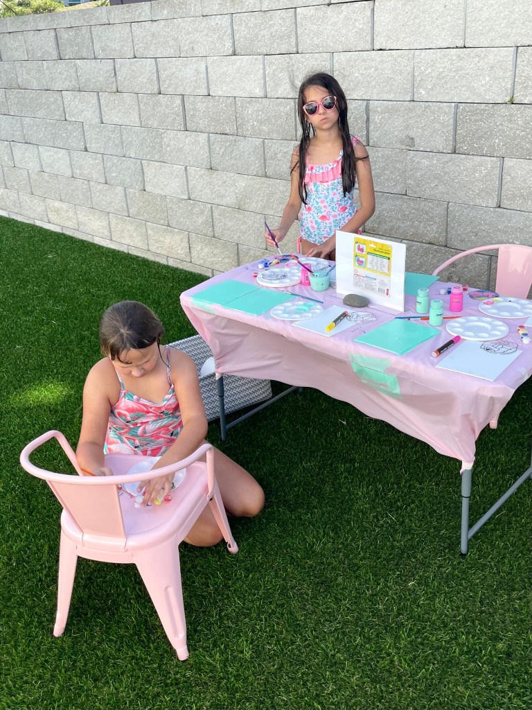 Two girls in swimsuits paint suncatchers at a party art station, surrounded by canvases drying with pastel backgrounds.