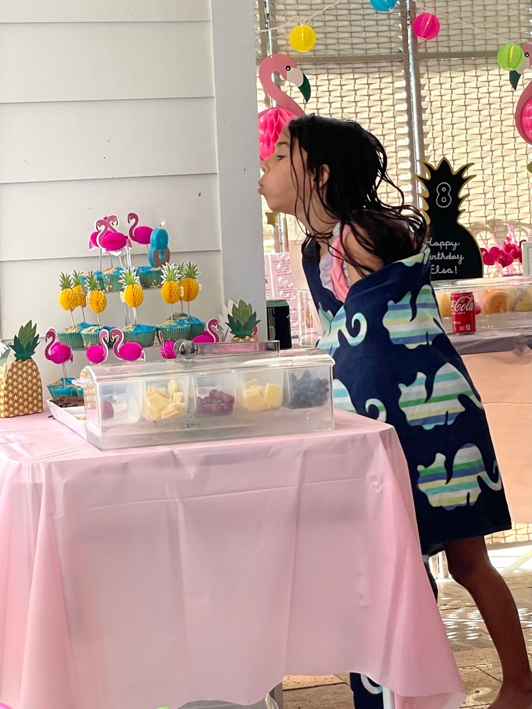 A young girl wrapped in a towel blows out the candle on a colorful cupcake stand decorated with flamingos and pineapples at her tropical birthday party.