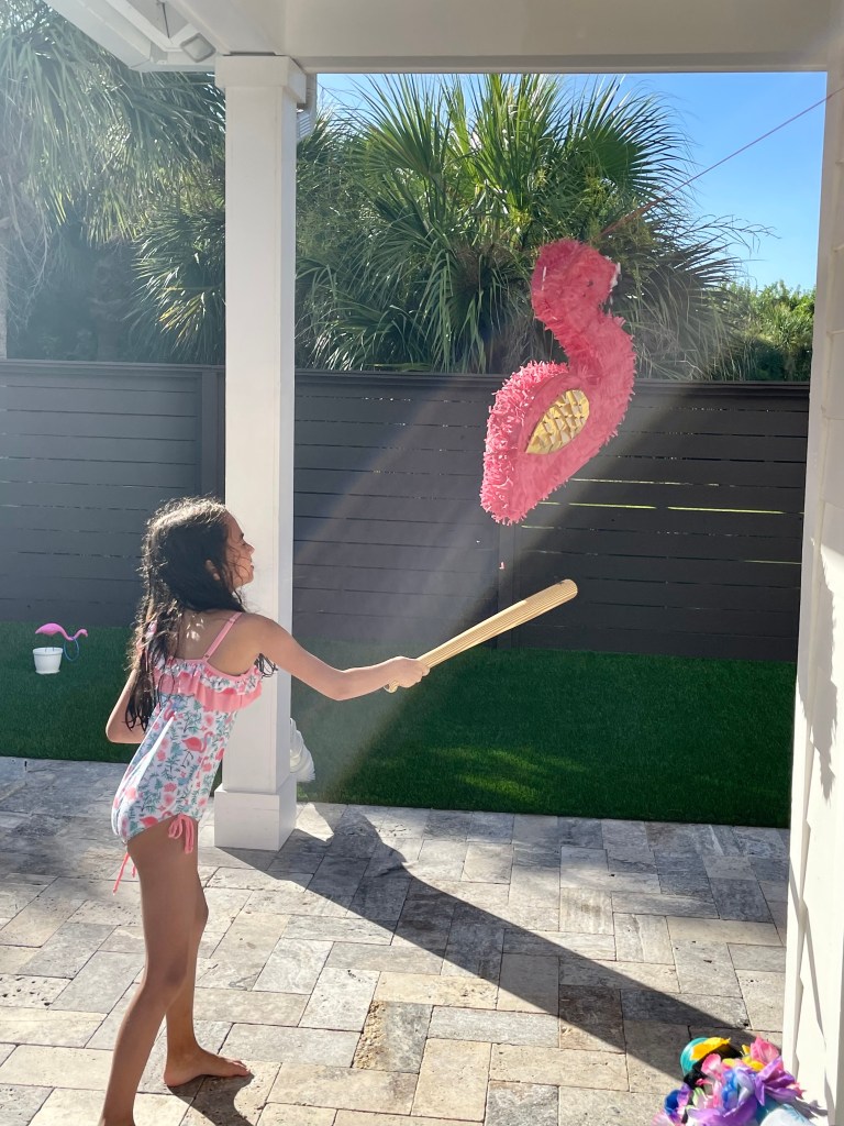 Girl in a flamingo swimsuit swings a bat at a pink flamingo piñata outside.