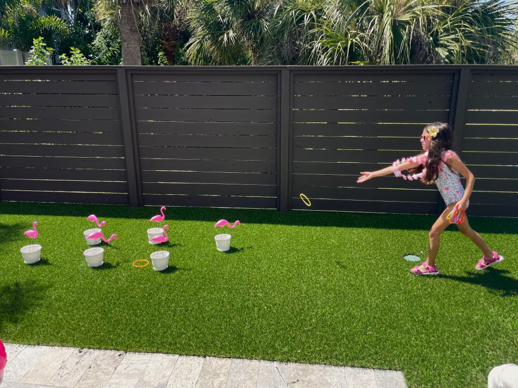 Girl in tropical-themed clothes throws a ring at a lawn flamingo during a backyard party game.