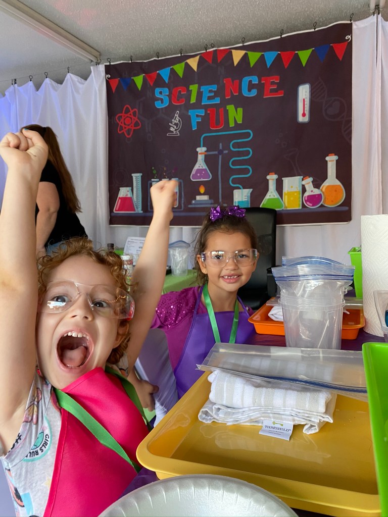 Two young girls wearing safety goggles and colorful aprons smile and raise their arms excitedly in front of a “Science Fun” banner, ready to begin the next activity.