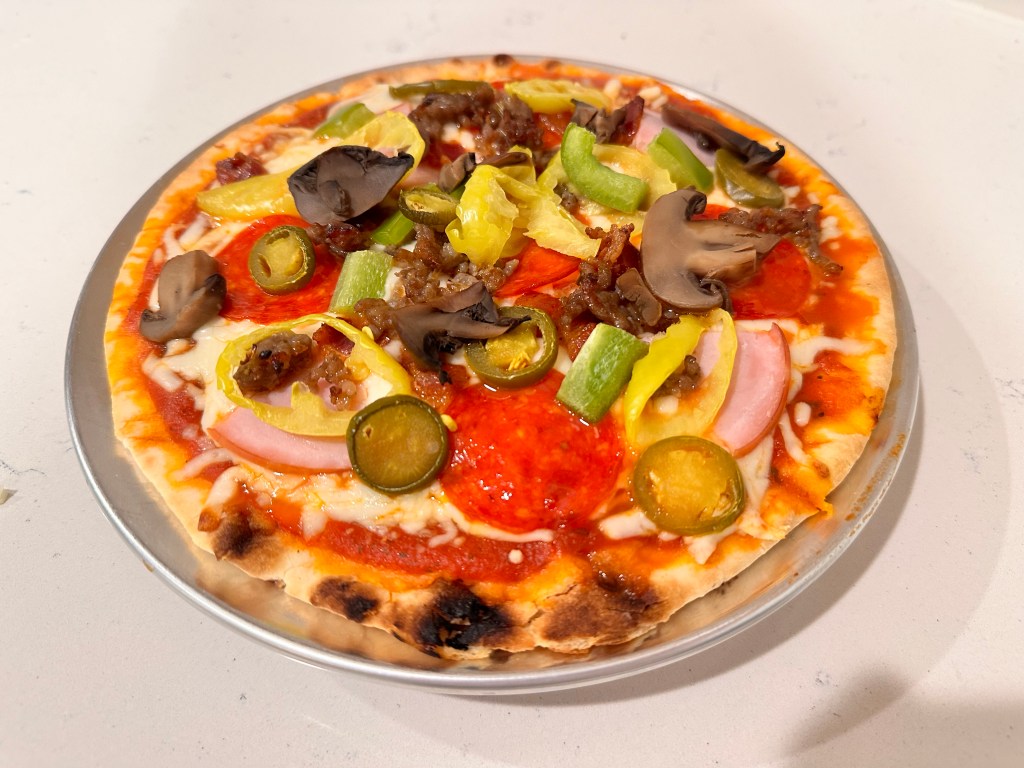 A baked personal pizza topped with pepperoni, mushrooms, jalapeños, and peppers on a silver pizza pan.