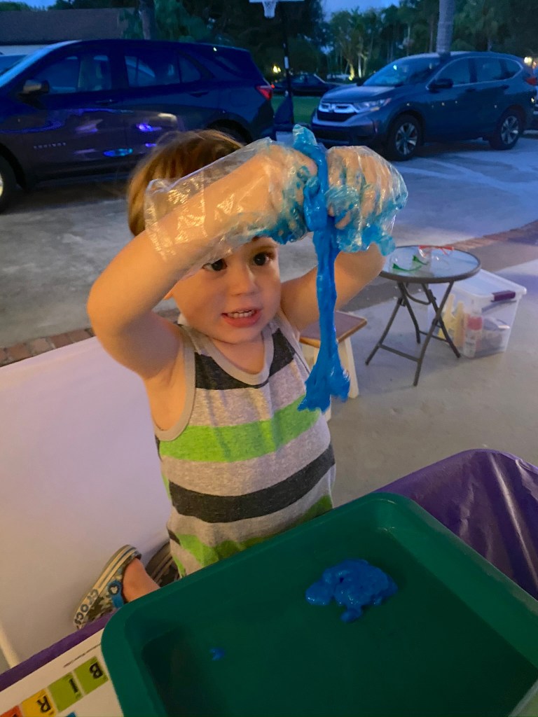 A young boy holds up bright blue slime, stretching it high above his head with a big smile.