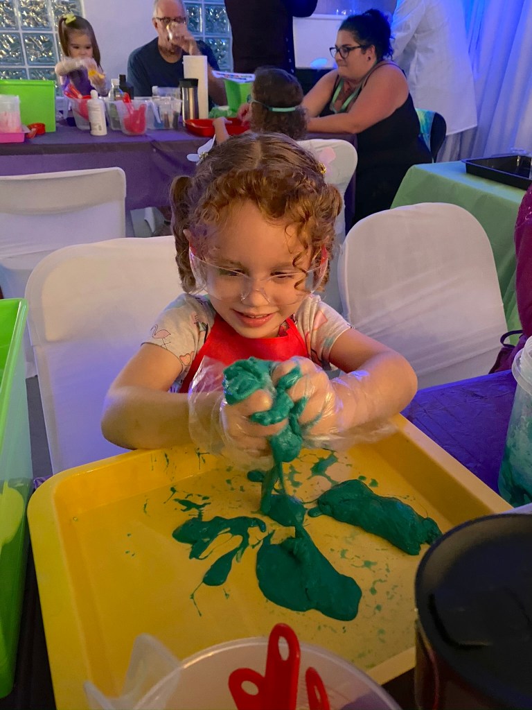 A smiling young girl wearing safety goggles squishes green slime between her hands over a yellow tray.