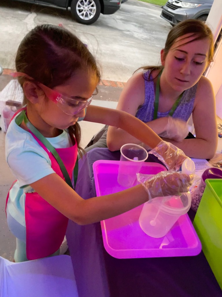 A young girl wearing safety goggles and gloves pours liquid into a container for making slime, with an adult supervising beside her.
