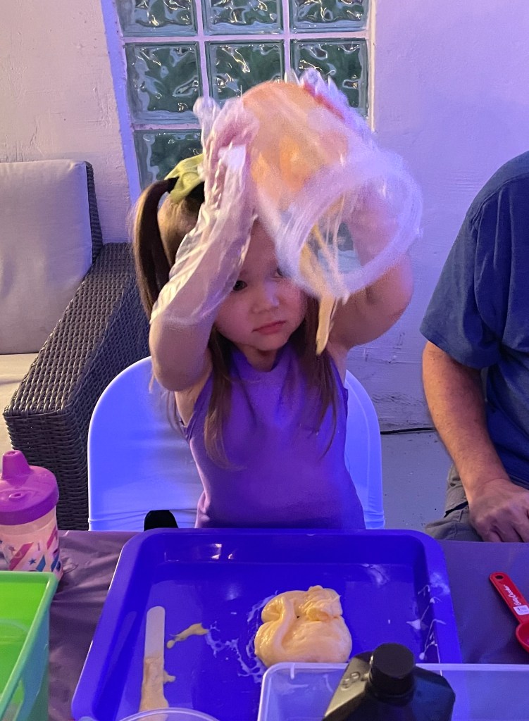 A young girl lifts a clear container above her head, letting orange slime drip onto the tray in front of her.