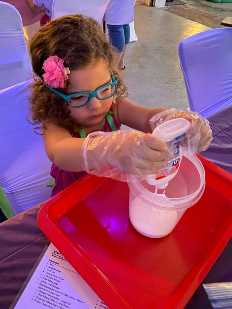 A young girl with glasses and gloves pours white glue from a bottle into a clear container for making slime.