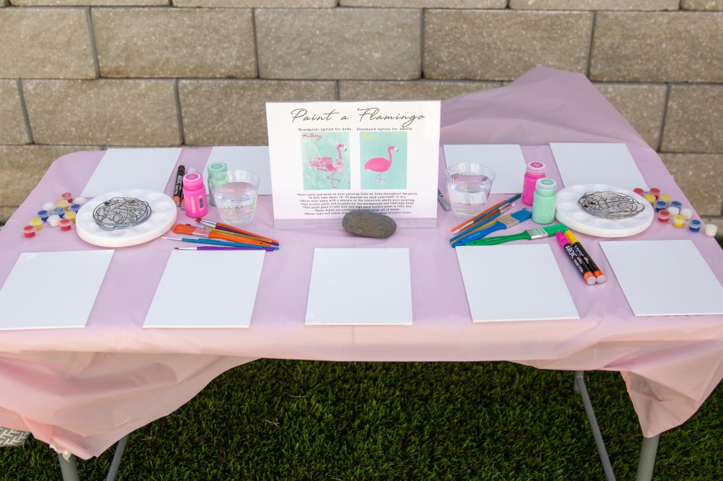 A pink outdoor table setup with canvases, brushes, paints, and a sign for painting flamingos at a kids’ birthday party.