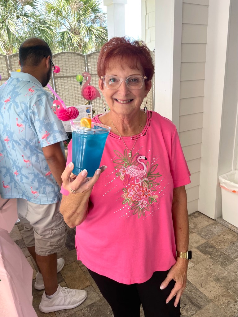 Smiling woman in a pink flamingo shirt holding a cup of blue tropical punch at a birthday party drink station.