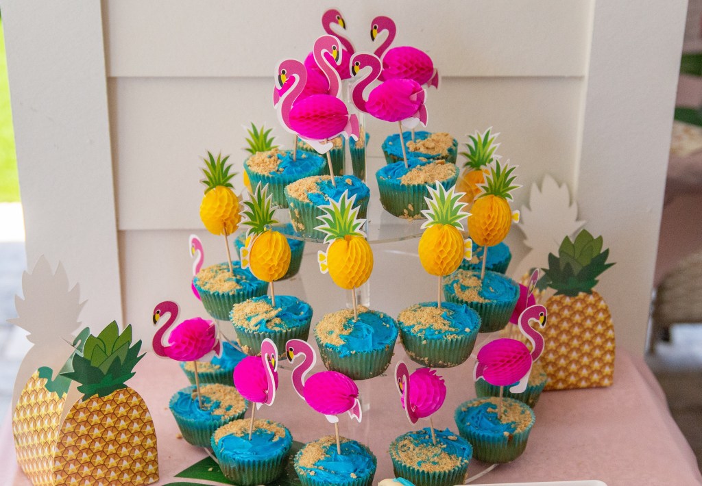 Flamingo and pineapple themed cupcakes with blue frosting and crushed graham cracker “sand” on a tiered cupcake stand