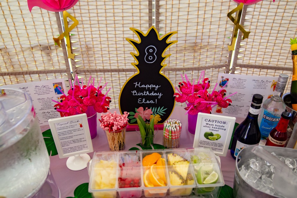 Close-up of a pineapple-shaped chalkboard reading “Happy Birthday Elsa” surrounded by tropical fruit garnishes and flamingo straws.