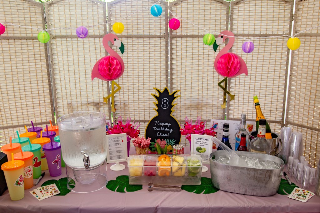 DIY tropical drink station with fruit, mix-ins, colorful cups, and flamingo decor for an outdoor pool party