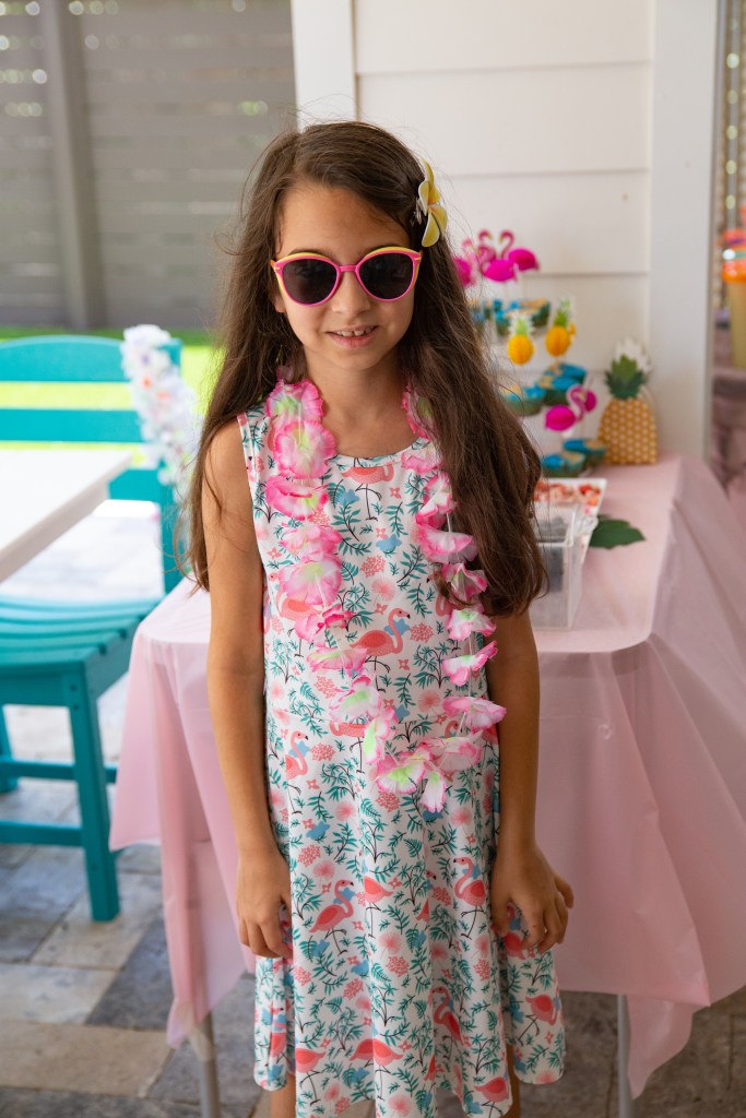 A girl in a flamingo-print dress and lei smiles in front of a pink-decorated party table at a tropical-themed birthday.