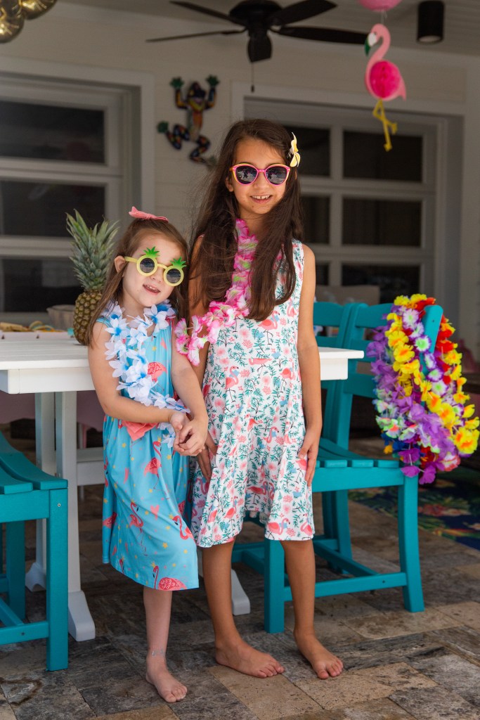Two young girls wearing flamingo-themed dresses and leis pose together at a tropical birthday party.