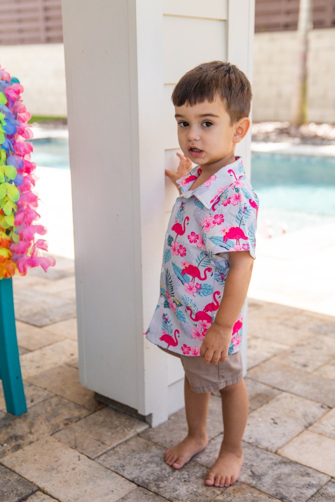 A young boy stands by a white patio column, wearing a button-up flamingo print shirt and tan shorts.