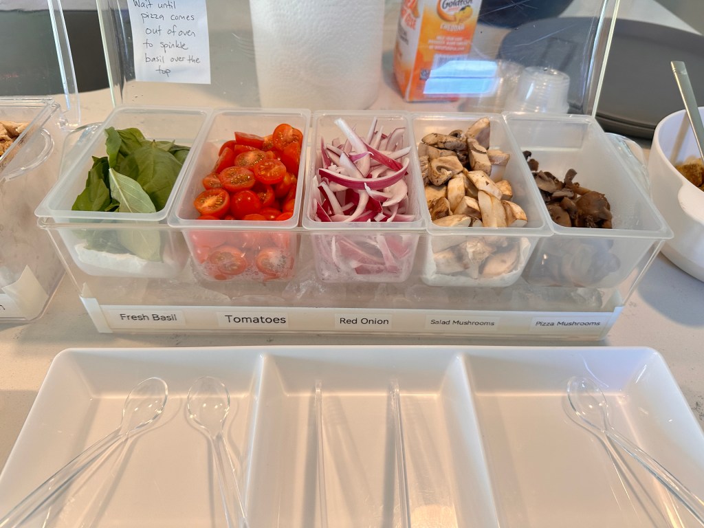 Pizza topping containers with fresh basil, grape tomatoes, red onion, and two types of mushrooms.