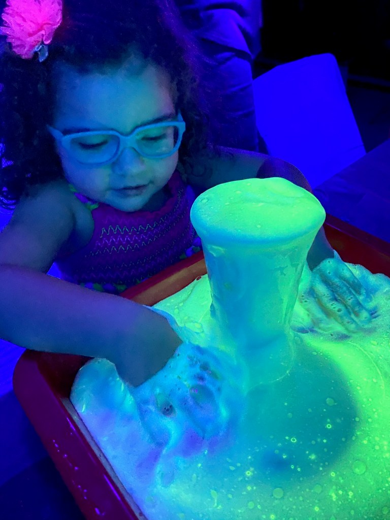 Close-up of a child with blue glasses playing in a tray of glowing green foam under UV light.