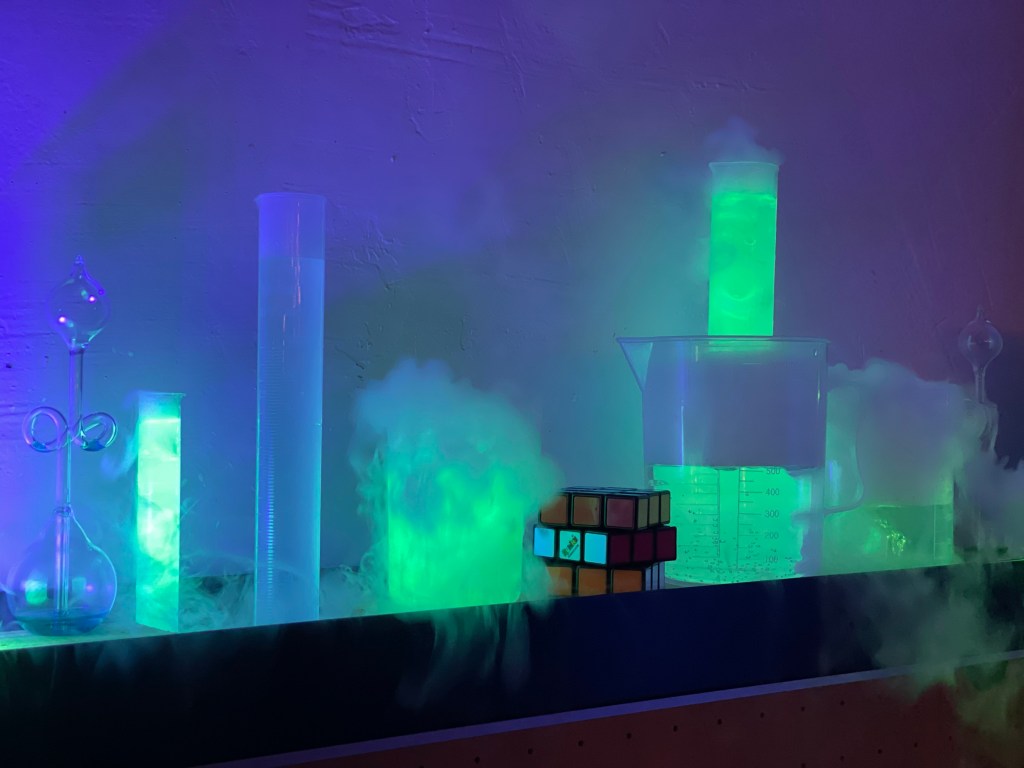 Glowing beakers and graduated cylinders filled with green liquid and surrounded by fog from dry ice, lit under blacklight.