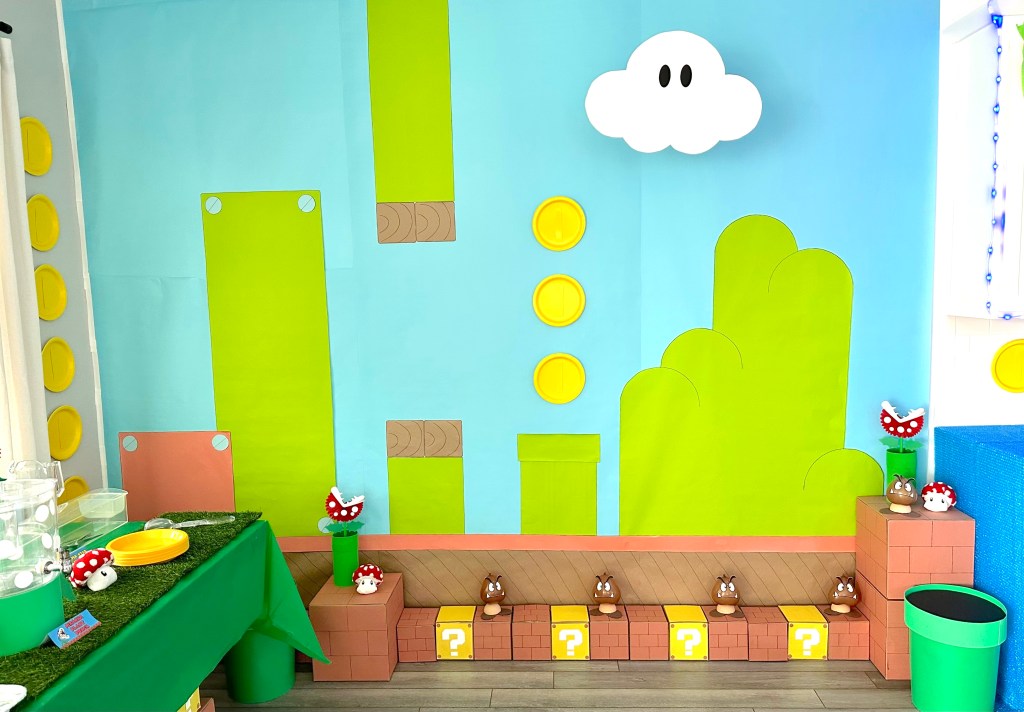Super Mario-themed Grass Land backdrop with blue sky, floating coins, green pipes, cardboard bricks, Piranha Plants, and Goombas for a birthday party decoration.
