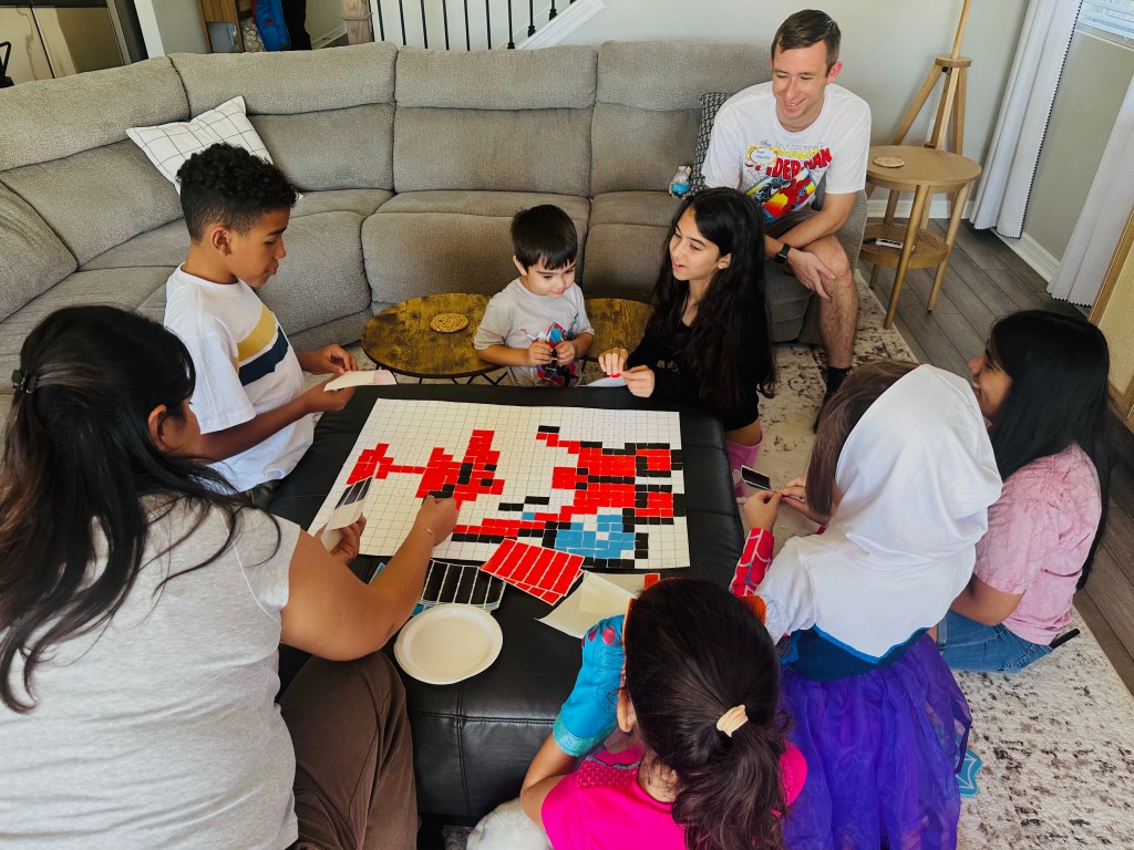 Group of children gathered around an ottoman working together to complete the Spider-Man sticker mosaic.