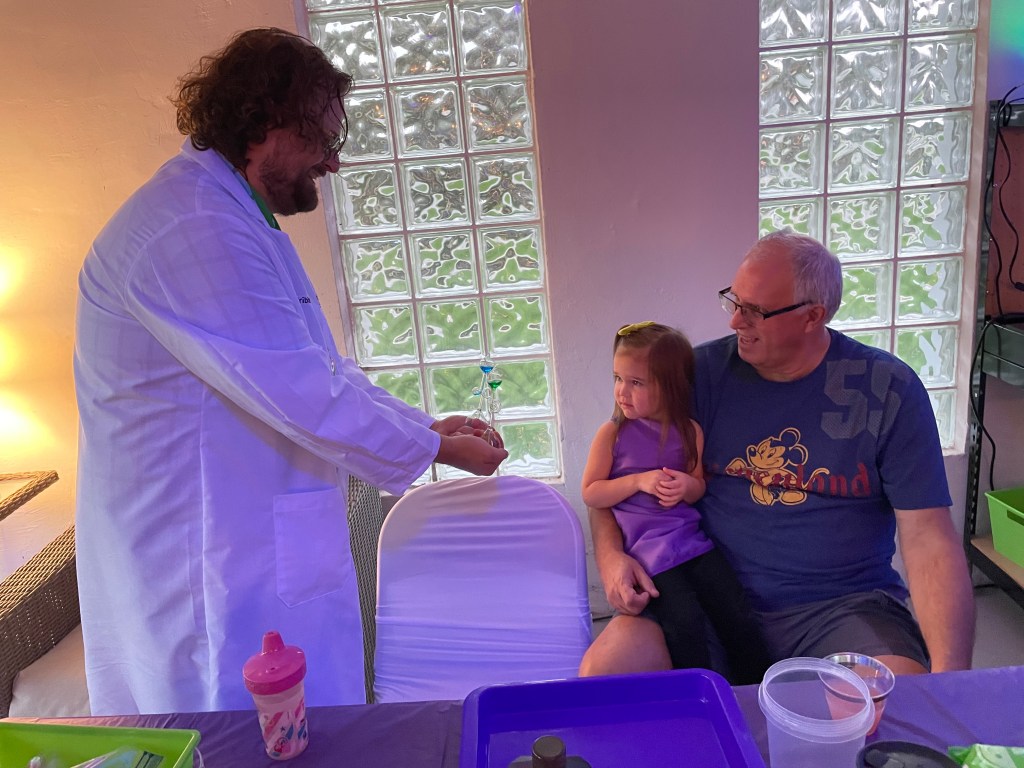 Child sitting on grandfather’s lap, both watching a man in a white lab coat hold a glass hand boiler in a science activity room.