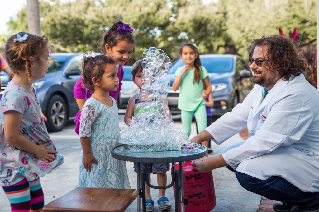 Man in lab coat creating a tall column of helium bubbles as children watch closely.